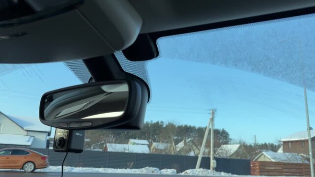 Car Interior Rearview Mirror with Dashcam in Winter. View of car rearview mirror and dashcam mounted on windshield. Blurred background shows snowy suburban street with houses and fences passing by.