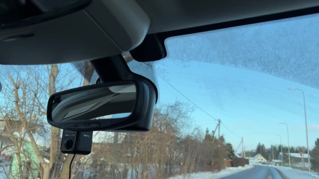 Rearview Mirror Reflection While Driving in Winter. Close-up of car rearview mirror reflecting rear window and road behind. Car is traveling through snowy village with houses and trees visible.