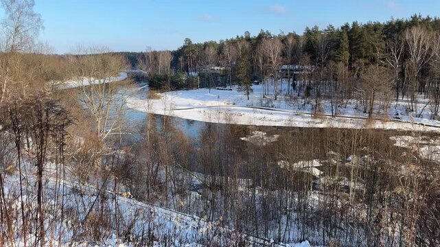 High Angle View of Winding River in Winter. Scenic wide shot overlooking river flowing through snowy landscape. Banks lined with bare trees and evergreens with distant houses nestled in forest.