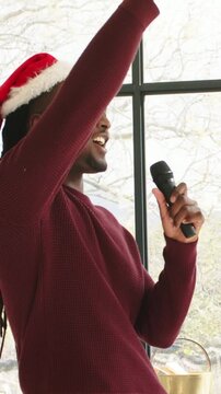 Vertical video: Holding wireless mic, man singing and gesturing for party by window, Santa hat
