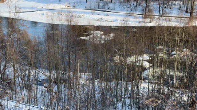 Winding River Flowing Through Snowy Winter Landscape. High-angle view of river winding through forest in winter. Banks covered in snow and ice with bare trees lining water under sunny sky.