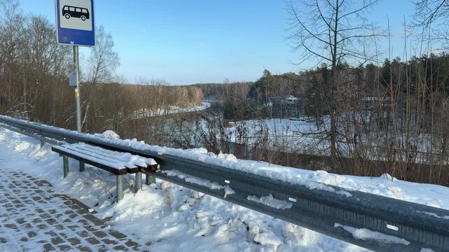 Bus Stop and Bench Overlooking Winter River Valley. Blue bus stop sign and wooden bench sit covered in snow on roadside. Background features scenic view of river valley and forest in winter.