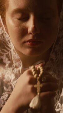 Vertical shot of young woman with red hair wearing white lace veil holding rosary beads near face while praying with eyes closed in studio under warm dim light, slow motion