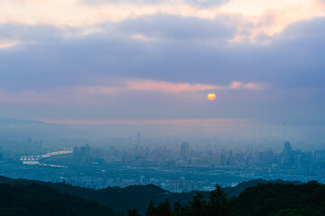 Fototapeta premium Sunrise over a misty cityscape from Dadao Mountain, Taiwan. The sun emerges from behind clouds, creating a dreamlike scene with layers of mountains and a vibrant orange sky.