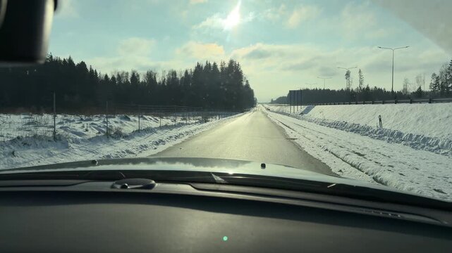 Driving Down Straight Winter Highway into Sun. Car travels along straight highway surrounded by snow-covered fields. Low winter sun shines brightly ahead illuminating wet road surface.