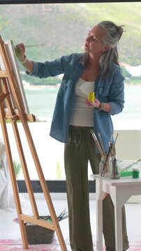 Vertical video: Picking yellow jar, woman painting at easel by window to create art, wearing denim