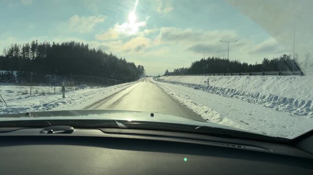 Driving on Sunlit Winter Highway. Car travels along straight highway flanked by high snow banks and pine forest. Bright winter sun shines directly ahead creating glare on wet asphalt road.