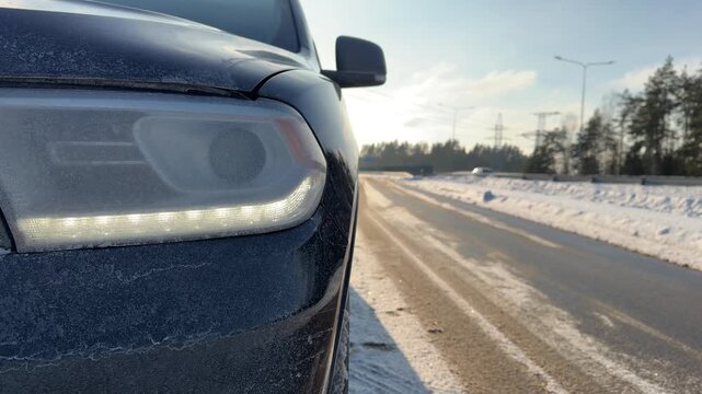 Frosted Car Headlight with LED Daytime Running Lights. Extreme close-up of car front headlight covered in frost with LED strips illuminated.Background shows sunny winter highway stretching in distance