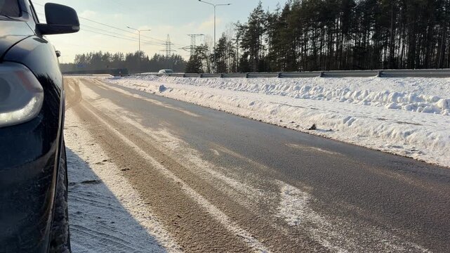 Roadside View of Winter Highway and Parked Car. Low-angle perspective looking down wet winter highway from side of parked black car. High snow banks line road and sun shines brightly in distance.