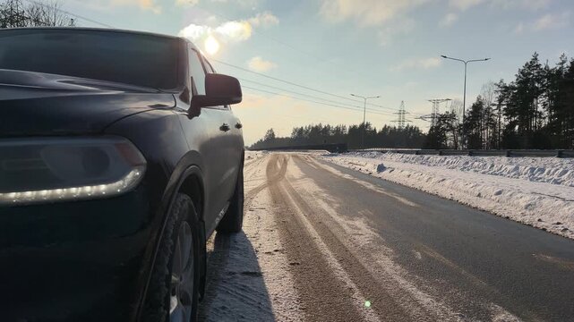 Low Angle of Parked SUV on Winter Highway with Sun. Low-angle shot captures black SUV parked on shoulder of winter highway. Camera pans from bright sun and clouds in blue sky down to vehicle and road.