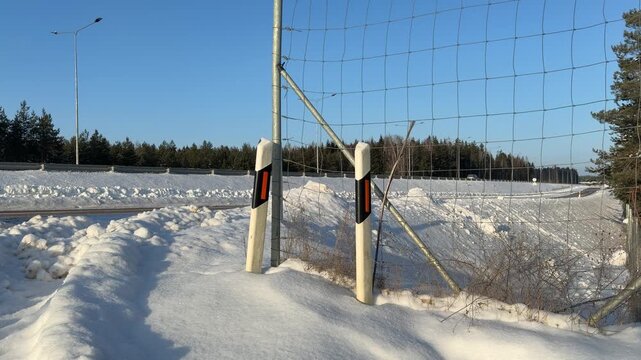 Road Reflectors and Fence Along Winter Highway. Two road marker posts stand in snow next to tall wire fence. Highway runs parallel in background with cars passing by on sunny winter day.