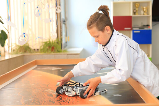 A focused boy in a white lab coat adjusts a robot on a table in a bright, creative STEM learning environment.