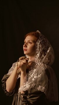 Vertical shot of teenage girl with red hair wearing floral lace hair scarf holding rosary beads in prayer while looking up with hope in low key lit studio against back background, slow motion