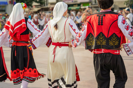 People in Bulgarian folk costumes holding hands dancing during cultural festival. Participants wearing traditional Bulgarian folk clothing dance in circle, public cultural festival in town center