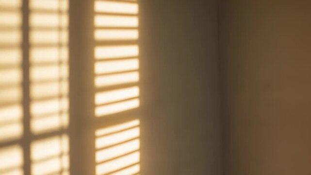 Warm sunlight filtering through blinds is illuminating an interior wall, casting long, geometric shadow patterns and creating a serene, inviting atmosphere with natural light