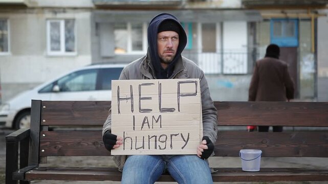 A homeless man in dirty clothes sits on a bench in a city park and asks for alms. Homeless man holds in his hands a paper with the inscription Help, I am hungry.