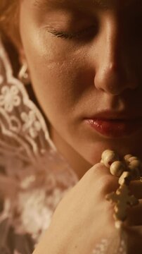 Vertical close up shot of young woman wearing white lace veil whispering prayer with eyes closed while holding rosary beads near face in studio under warm low key light, slow motion