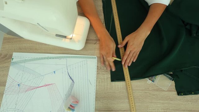 A person marks measurements on dark green fabric using a wooden ruler and yellow chalk, beside a sewing machine and pattern sheet on a wooden table.
