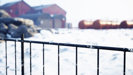A black fence stands in the foreground of a serene snowy landscape. Bright sunlight shines on old red buildings in the background, creating a peaceful winter scene. © icetray