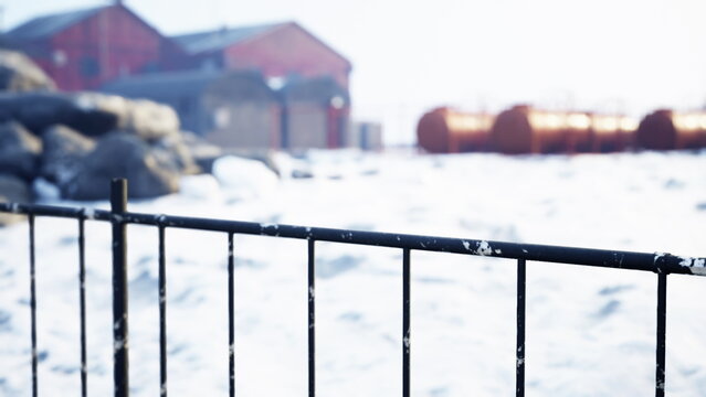 A black fence stands in the foreground of a serene snowy landscape. Bright sunlight shines on old red buildings in the background, creating a peaceful winter scene.