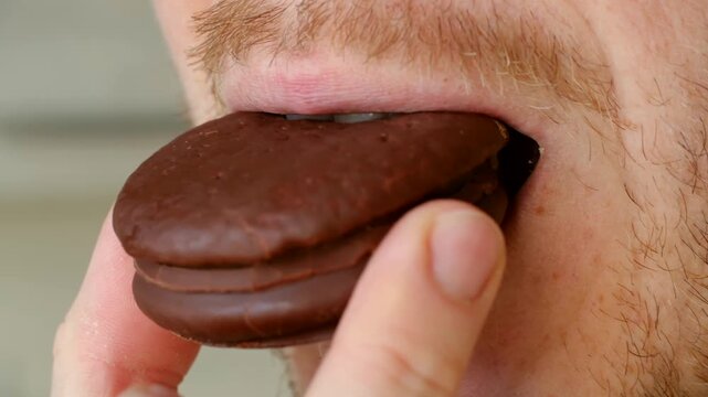 Close up man with light beard eating chocolate covered sandwich cookie, showing details of his mouth, lips, and teeth as he takes bite, chews with satisfaction. Man eating chocolate biscuit cookie