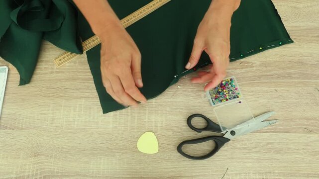 A person pins a folded hem on dark green fabric using colorful straight pins, with scissors, a ruler, and pattern piece nearby on a wooden table.