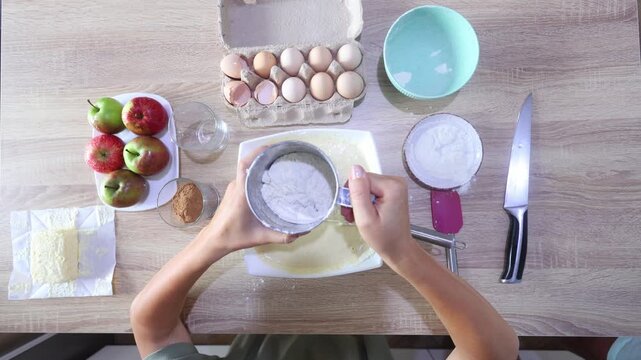 Top-down view: hands sift flour into a bowl of wet batter, then whisk, surrounded by apples, eggs, milk, cinnamon, and butter on a clean kitchen counter.