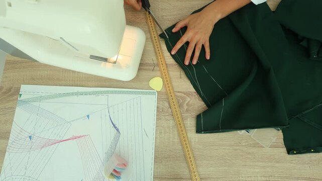 A person marks measurements on dark green fabric using a wooden ruler and yellow chalk, beside a sewing machine and pattern sheet on a wooden table.