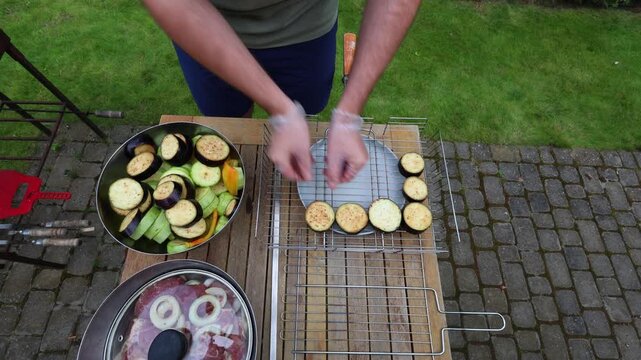 A man wearing gloves arranges sliced eggplant and zucchini on a grill basket and plate, preparing vegetables for outdoor grilling.