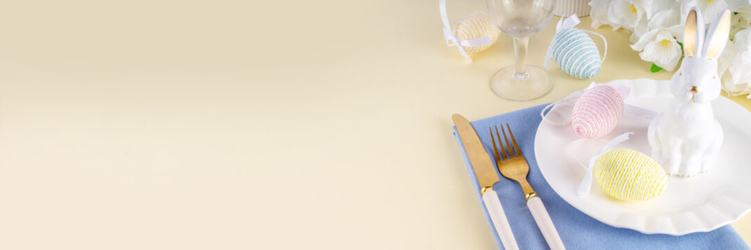 Cute Easter table setting with plate, cutlery set bunny, Easter egg, napkin and spring flowers on pastel colored yellow background, copy space, Easter brunch on dinner invitation, cafes menu layout
