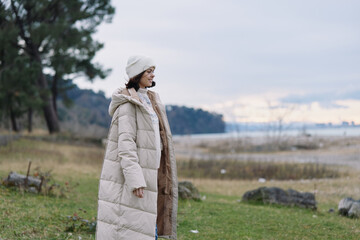 Fototapeta premium Woman in a long beige puffer coat and hat standing near a lake, autumn park landscape, calm sky, distant trees, soft light, outdoor fashion and travel mood.