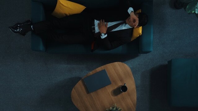 Top down aerial view of stressful boss look at decrease sales from laptop and lies on sofa. Tired manager working laptop while feeling stress. Skilled businessman having headache at work. Directorate.