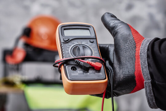 Electrician wearing protective gloves holding digital multimeter for electrical testing and diagnostics. Concept of professional electrical service, voltage measurement and equipment inspection.