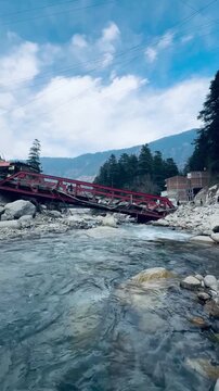 Collapsed red iron bridge over a mountain river after severe flooding, showing natural disaster damage in a Himalayan town with rocky debris and strong water current.