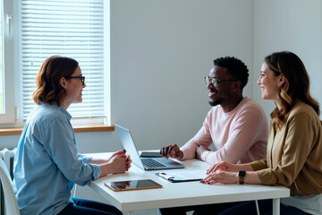 Young woman interviewing Black young man and young woman sitting at table using laptop and tablet smiling and engaging in conversation
