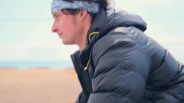 White man cycling on coastal trail with black puffer jacket -  riding bicycle along sandy path -  bare trees and windy sea horizon -  steady cadence and solitary reflective mood