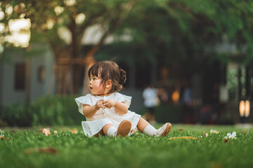 Adorable little Asian girl in a white dress sitting on a green lawn and playing with white flowers in a park during summer