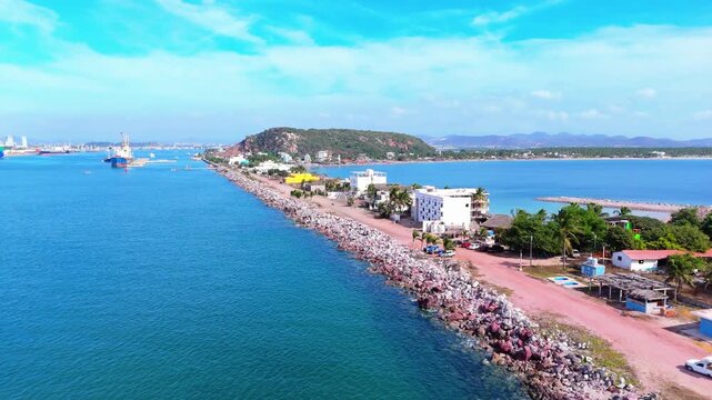 Vista a&eacute;rea costera mostrando un rompeolas de piedra junto a una zona urbana frente al oc&eacute;ano Pac&iacute;fico. Se observan edificios residenciales, palmeras, una carretera costera y el mar en calma bajo un c