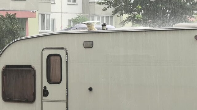 Rain pours down on a motorhome, soaking its roof and side. The scene unfolds in a calm neighborhood as storm clouds gather overhead. Nearby buildings frame the backdrop.