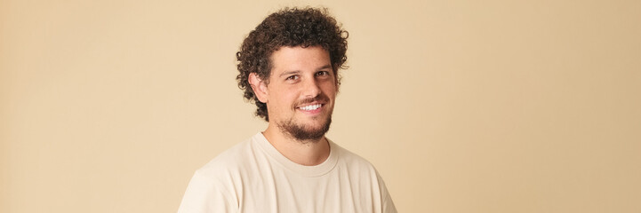 Fototapeta premium Happy guy with curly hair dressed in beige t-shirt laughing looking at camera isolated on beige background in studio. Panoramic