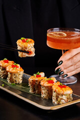 A lifestyle food photography shot featuring a woman in a black blazer holding a chilled orange cocktail in a coupe glass. picking up a gourmet sushi roll with chopsticks