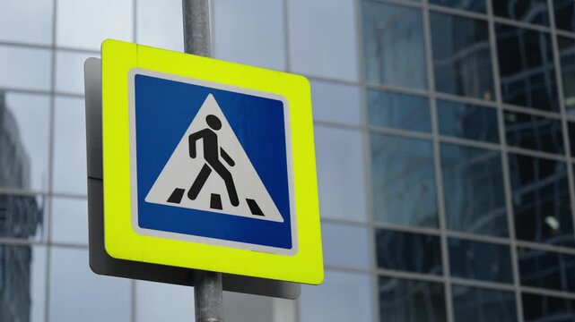 Close-up of a pedestrian crossing traffic sign with a bright yellow reflective border and blue triangle symbol on a pole, against a modern glass building facade in urban setting