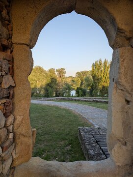 Curved stone archway frames a tranquil outdoor scene with a winding cobblestone path, green grass, and a lush riverside forest under a clear blue sky. Warm light enhances the textures of the old mason