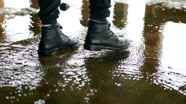 Close-up of waterproof boots stepping into a puddle, causing concentric water rings. Stomping through a puddle, capturing the moment of impact and resulting water spray. Feet in dark boots walking