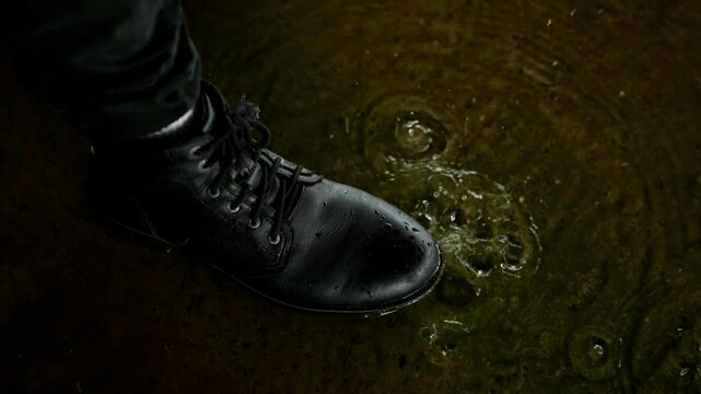 Black boots stand at the edge of a murky puddle, reflecting overhead light in the damp, urban setting. Close-up on dark trousers and lace-up boots near rippling water where raindrops create small spla
