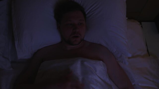 High angle view of terrified man abruptly waking up in his bed at night from bad dream, breathing heavily with scared expression and clutching his chest in slow motion, experiencing panic attack