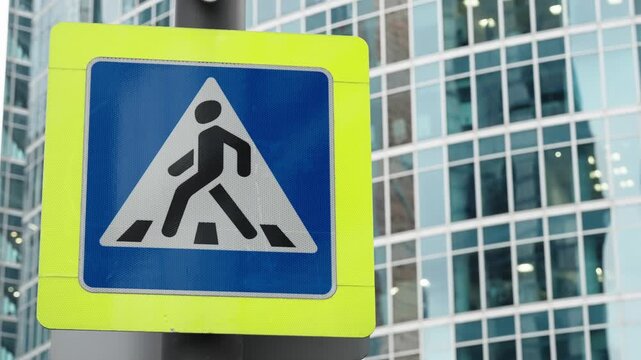 High-visibility pedestrian crossing sign with blue triangular symbol and zebra lines on a pole, set before modern glass skyscraper facades in a dense, contemporary urban cityscape