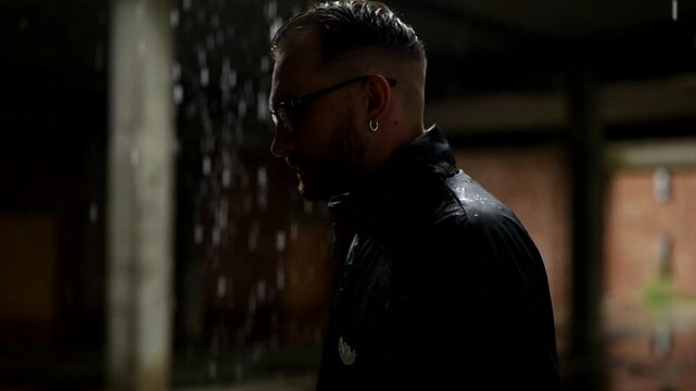 Man in black attire standing in a decaying, damp interior as water streams from the ceiling. Dripping water illuminates a dark, unfinished concrete space where a suited figure stands. Depressed man in