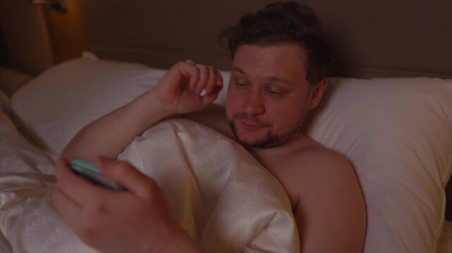 Tired caucasian man with a smartphone in bed at night, rubbing his eyes from fatigue while doomscrolling on social media instead of sleeping, suffering from insomnia, phone addiction