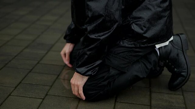 Man in black grief-stricken raincoat kneeling on wet, dark paving stones, head bowed. Solitary figure in glossy black attire against a muted blue background, bowed in rain.